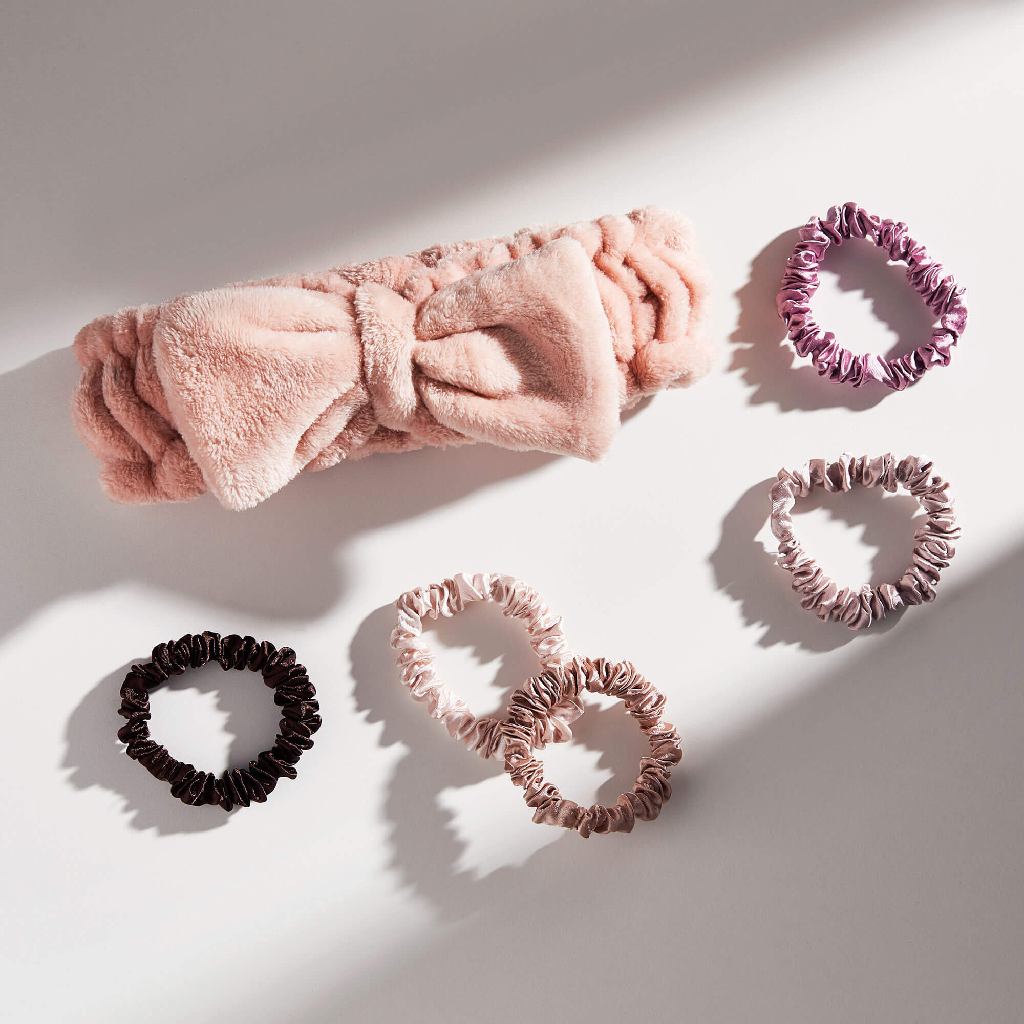 A pink plush headband with a bow and five scrunchies in shades of purple and brown are arranged on a white surface with shadows.