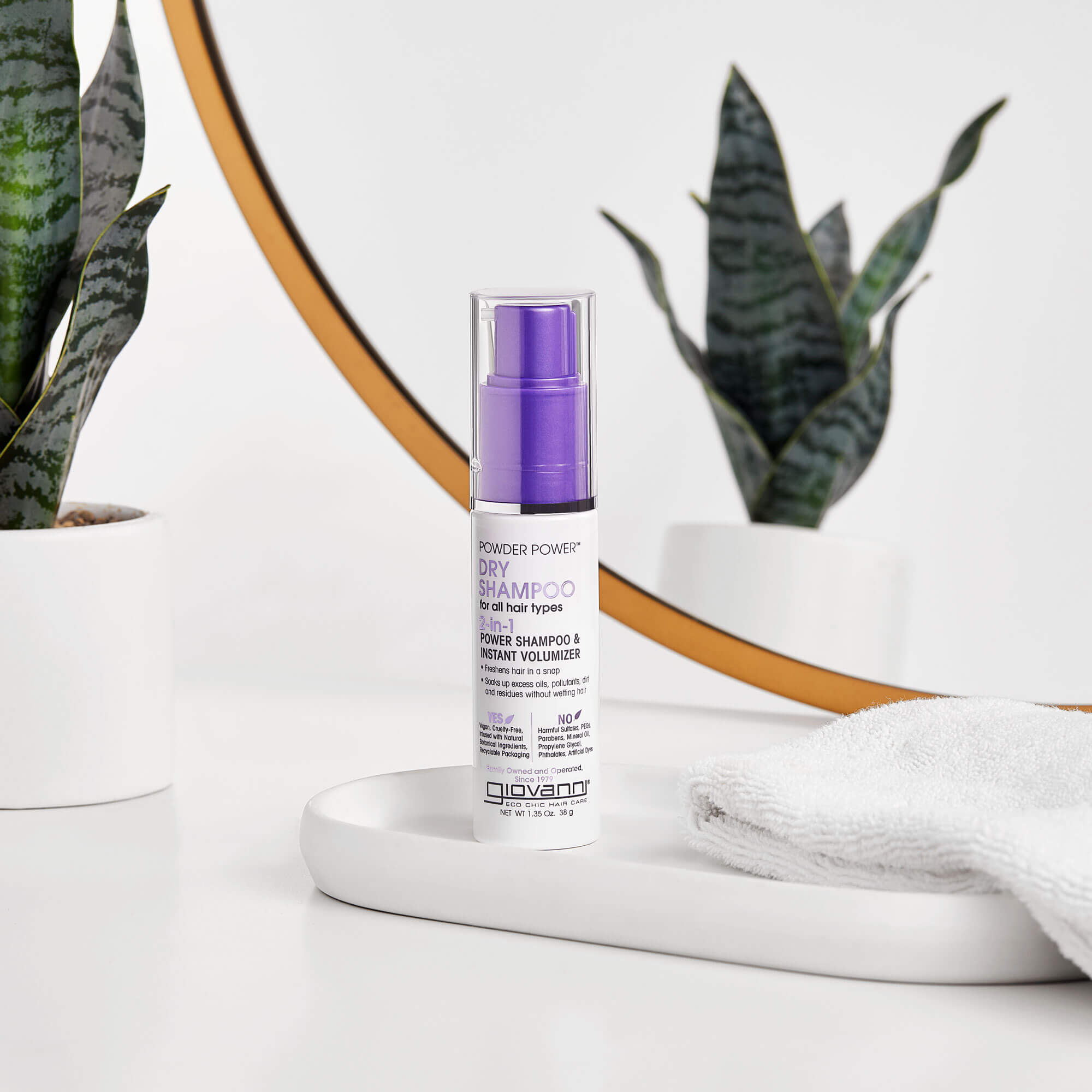 A bottle of Giovanni Powder Power Dry Shampoo with a purple cap sits on a white tray next to a folded towel, with potted plants and a round mirror in the background.