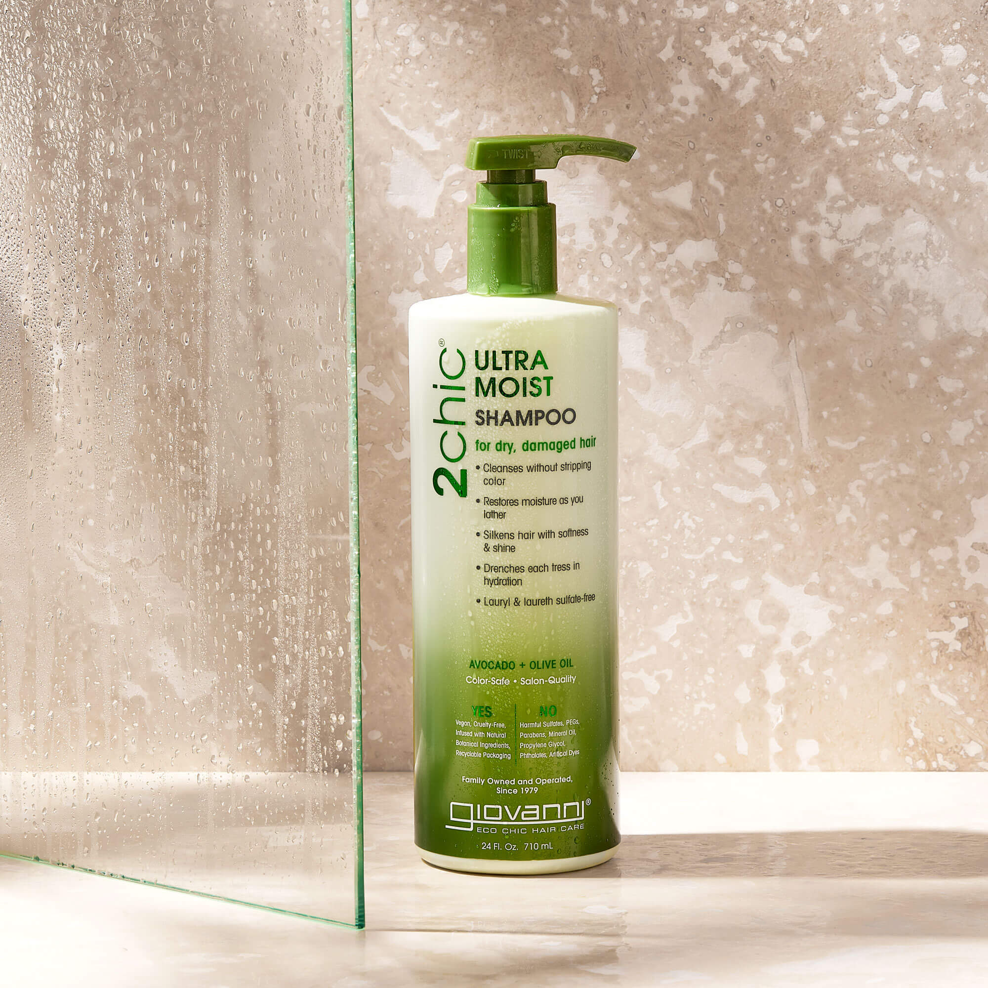 A green bottle of Giovanni 2chic Ultra Moist Shampoo with a pump sits on a wet beige surface near a glass shower panel, surrounded by water droplets.
