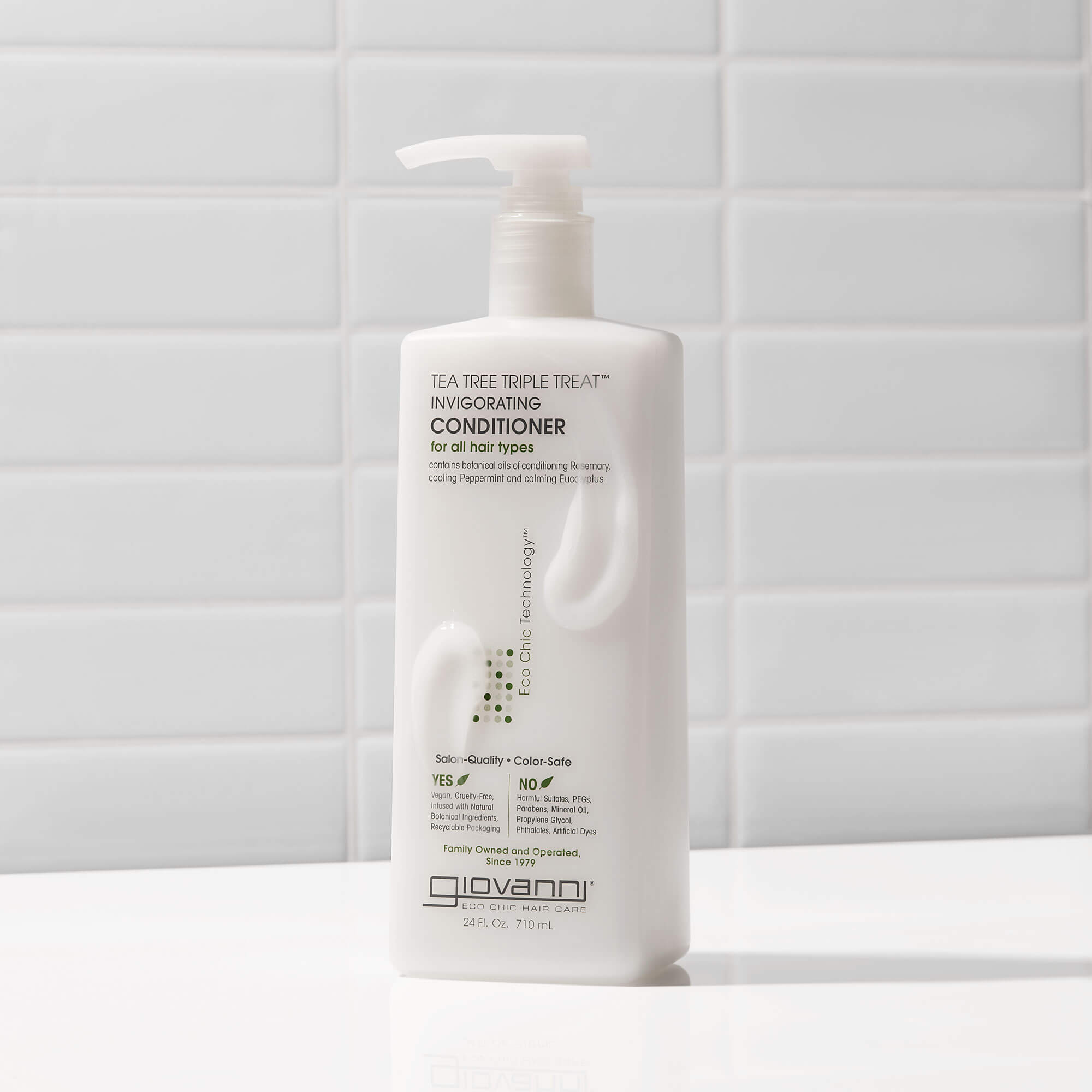 A white pump bottle of Giovanni Tea Tree Triple Treat Invigorating Conditioner stands on a white surface against a white tiled background.