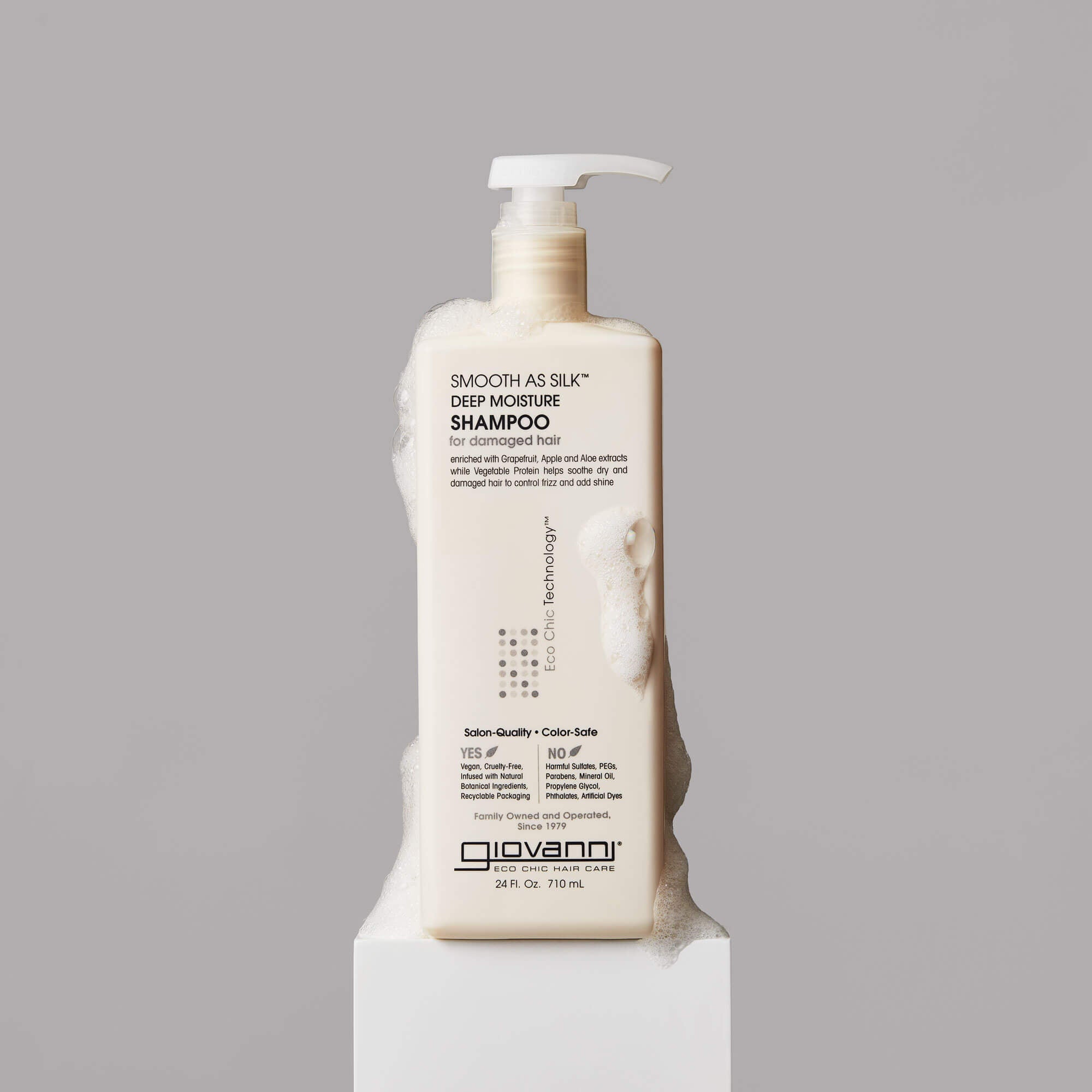 A beige bottle of Giovanni Smooth as Silk Deep Moisture Shampoo with a pump top, covered in foamy bubbles, stands on a white pedestal against a gray background.
