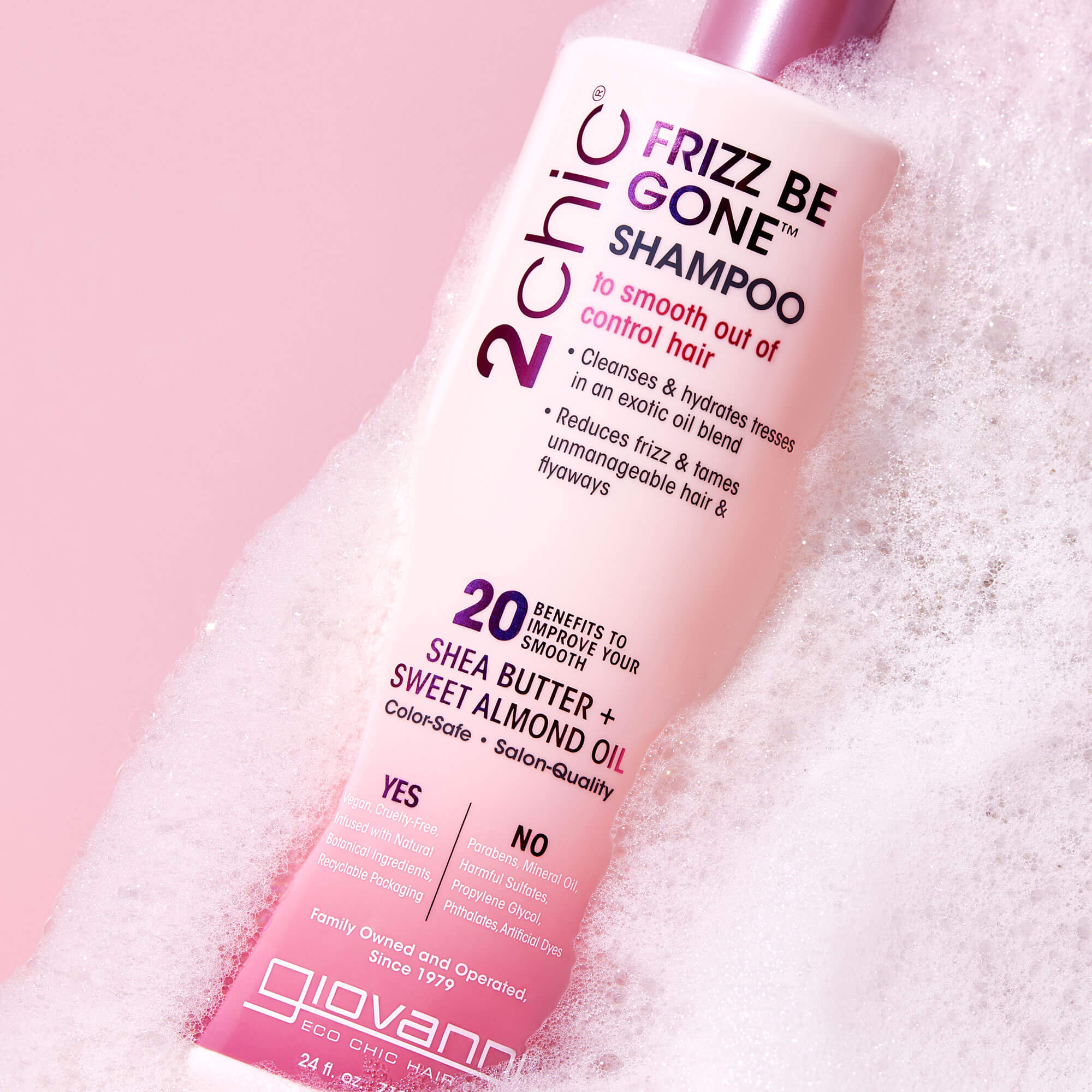 A bottle of Giovanni 2chic Frizz Be Gone Shampoo rests in pink-tinted bubbles. The label highlights frizz control benefits and key ingredients like shea butter and sweet almond oil.