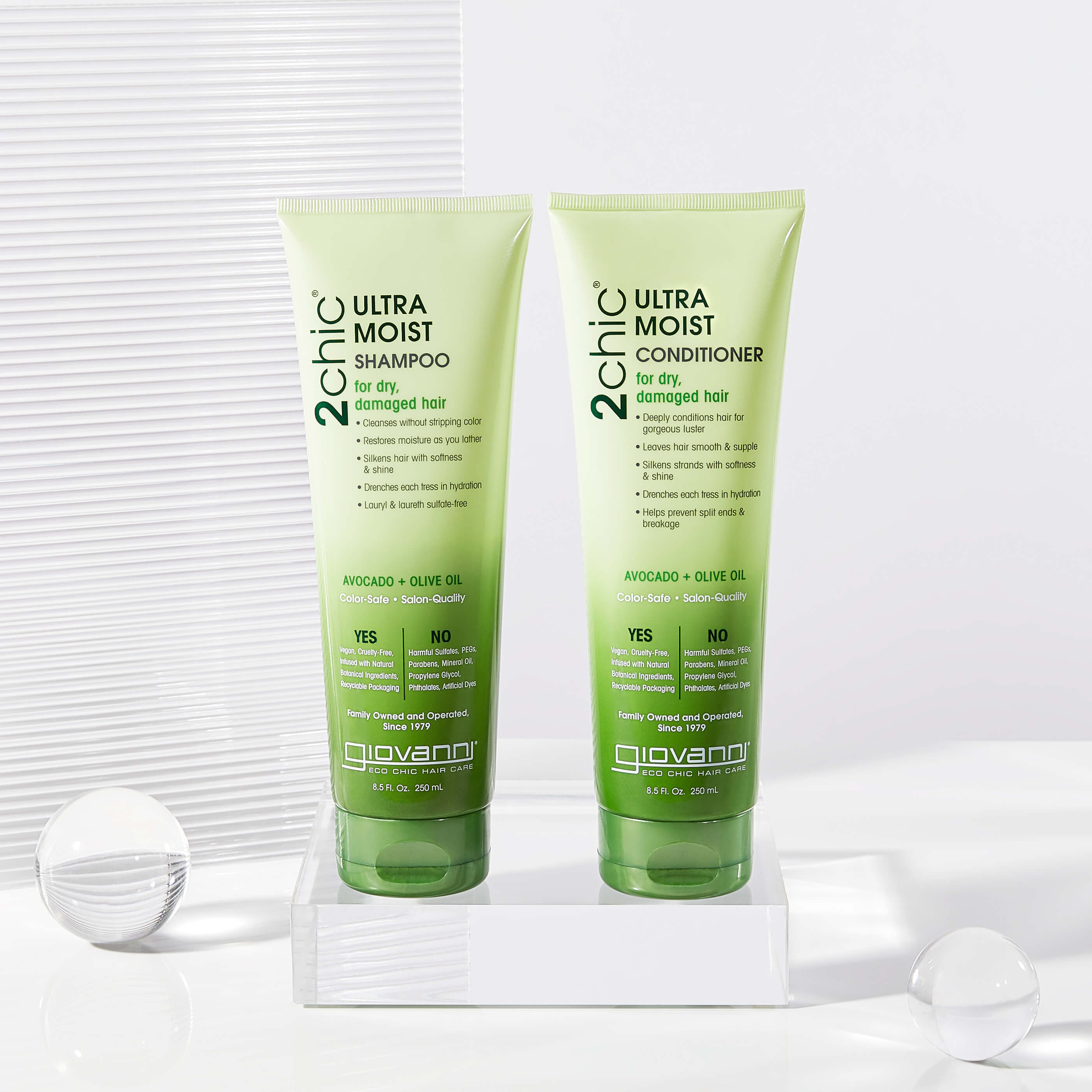 Two green bottles of Giovanni 2chic Ultra Moist Shampoo and Conditioner for dry, damaged hair are displayed on a white surface with glass spheres and soft lighting in the background.