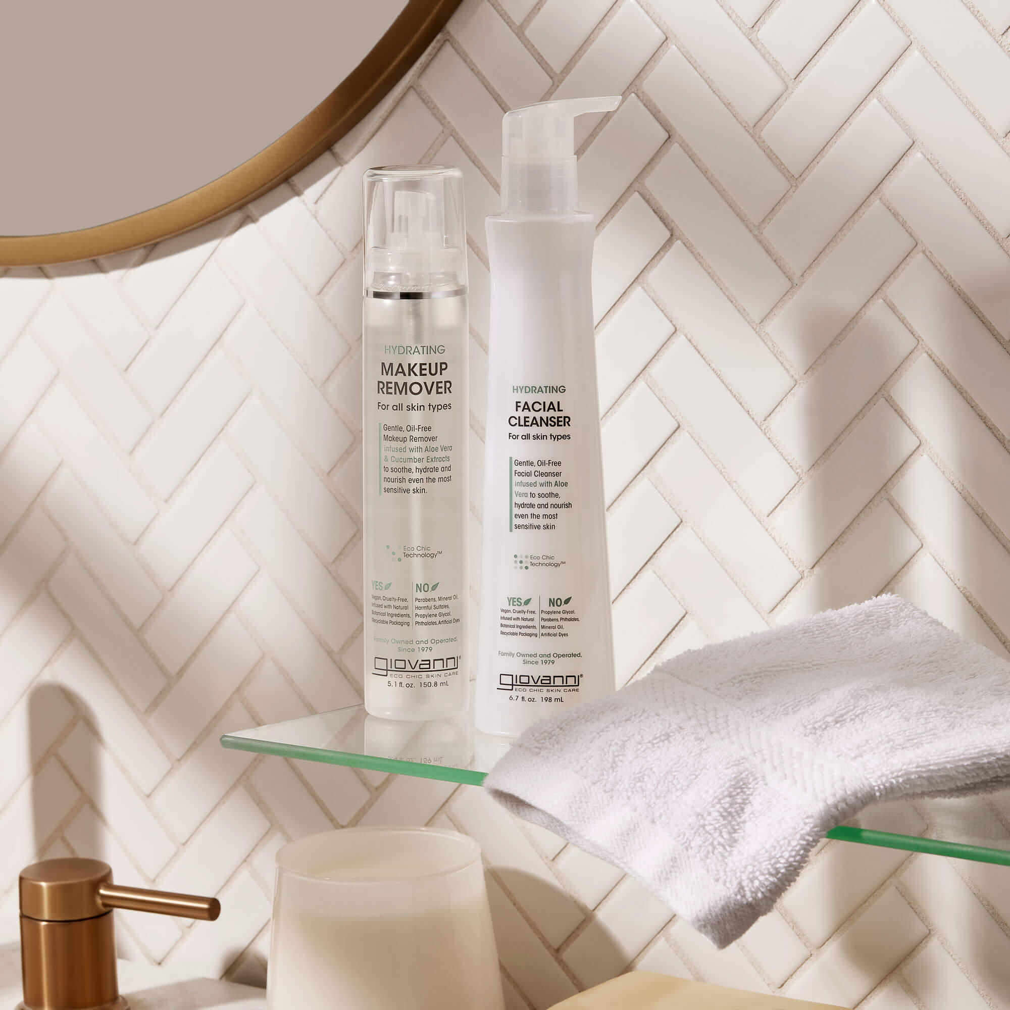 Two clear bottles labeled Makeup Remover and Facial Cleanser stand on a glass shelf with a white towel, against a white herringbone tile wall and a round mirror.