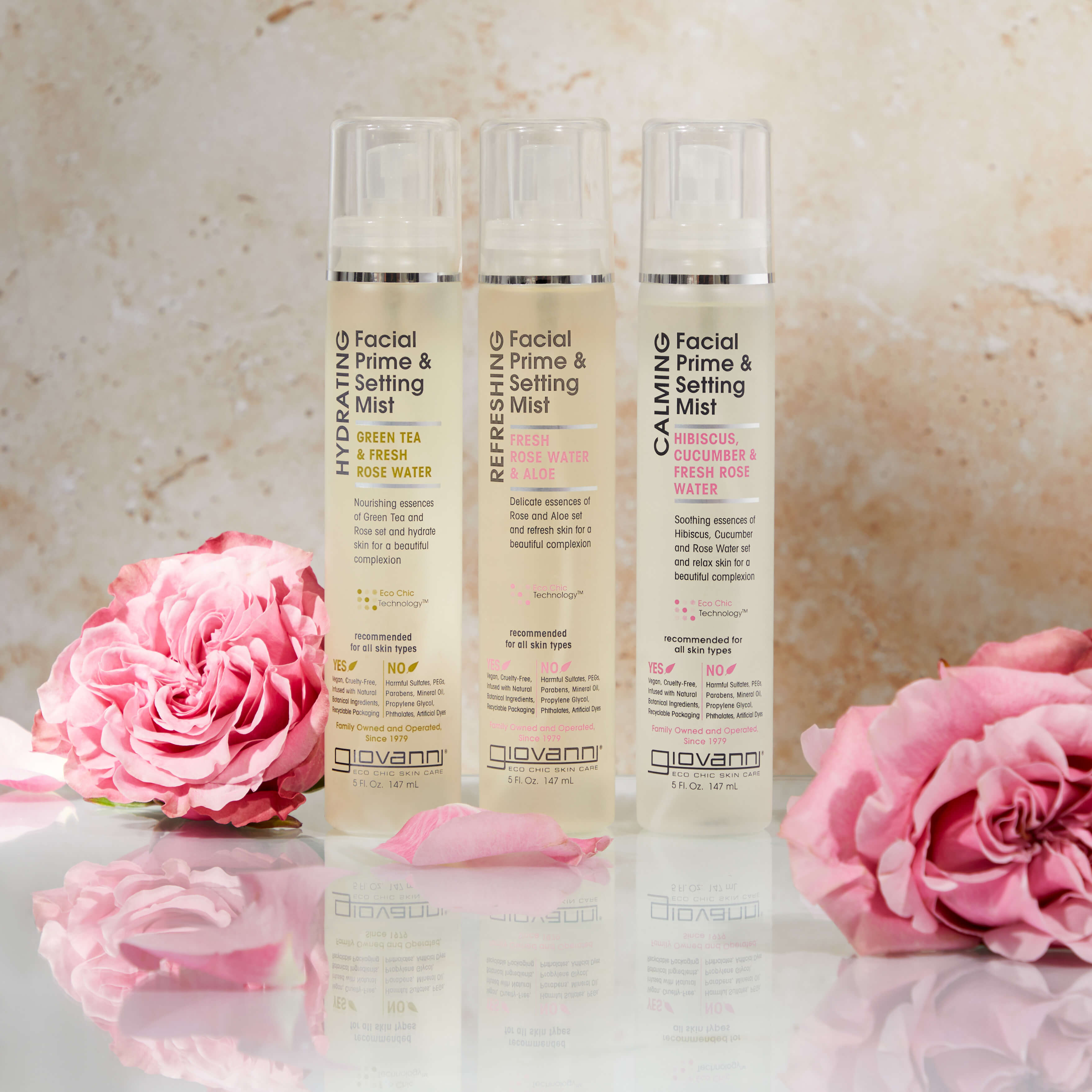 Three bottles of Giovanni facial prime and setting mist stand upright between two pink roses, against a light beige marble background. The bottles are labeled Hydrating, Refresh, and Calming.