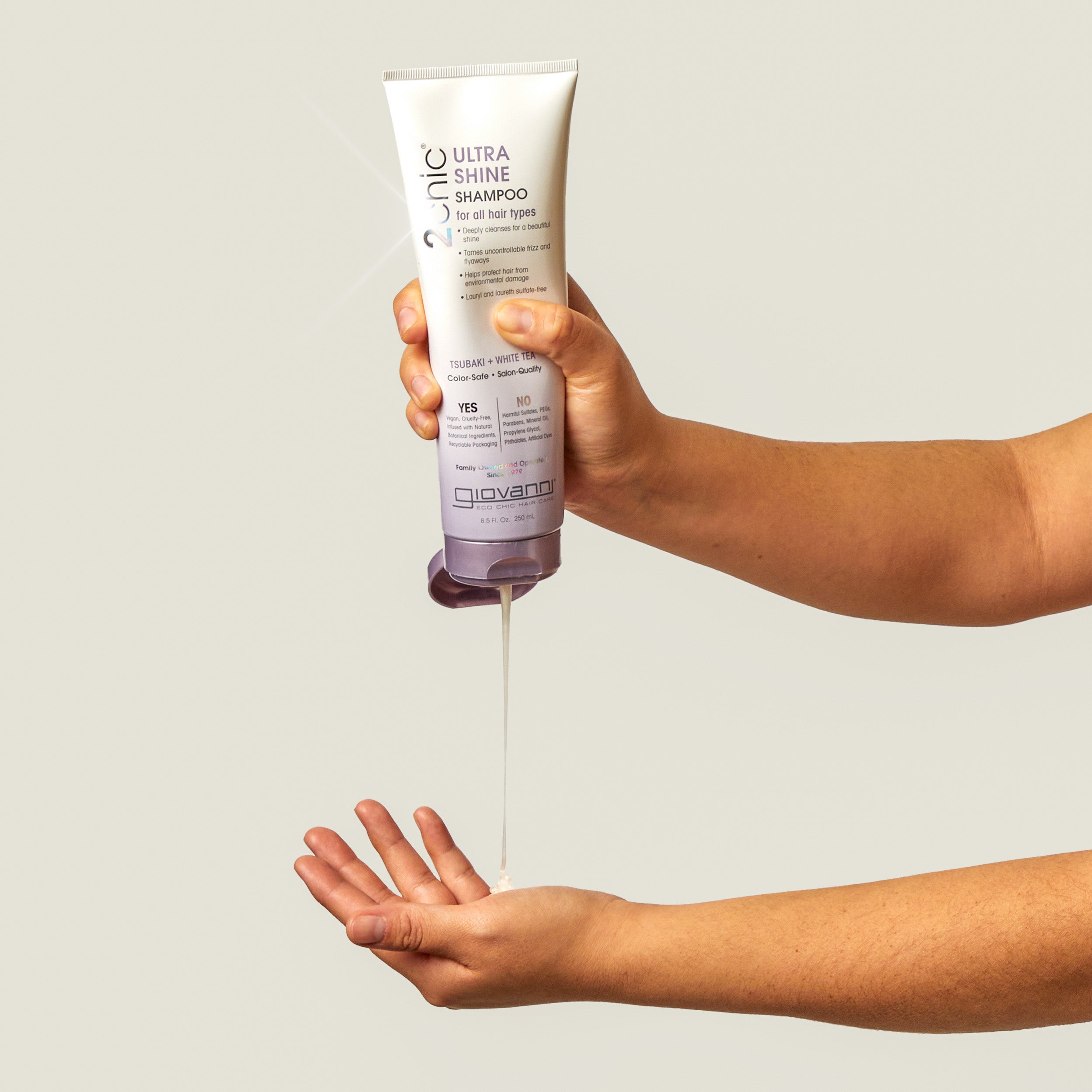Hand holding a bottle of Giovanni Ultra Shine Shampoo with liquid being poured onto another hand against a neutral background.