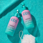 Two blue and purple pump bottles of Giovanni Cosmetics ECO CHIC® KIDS 2 IN 1 SHAMPOO & CONDITIONER sit on a turquoise towel next to a white bath pouf, featuring fun, colorful children’s haircare labels.