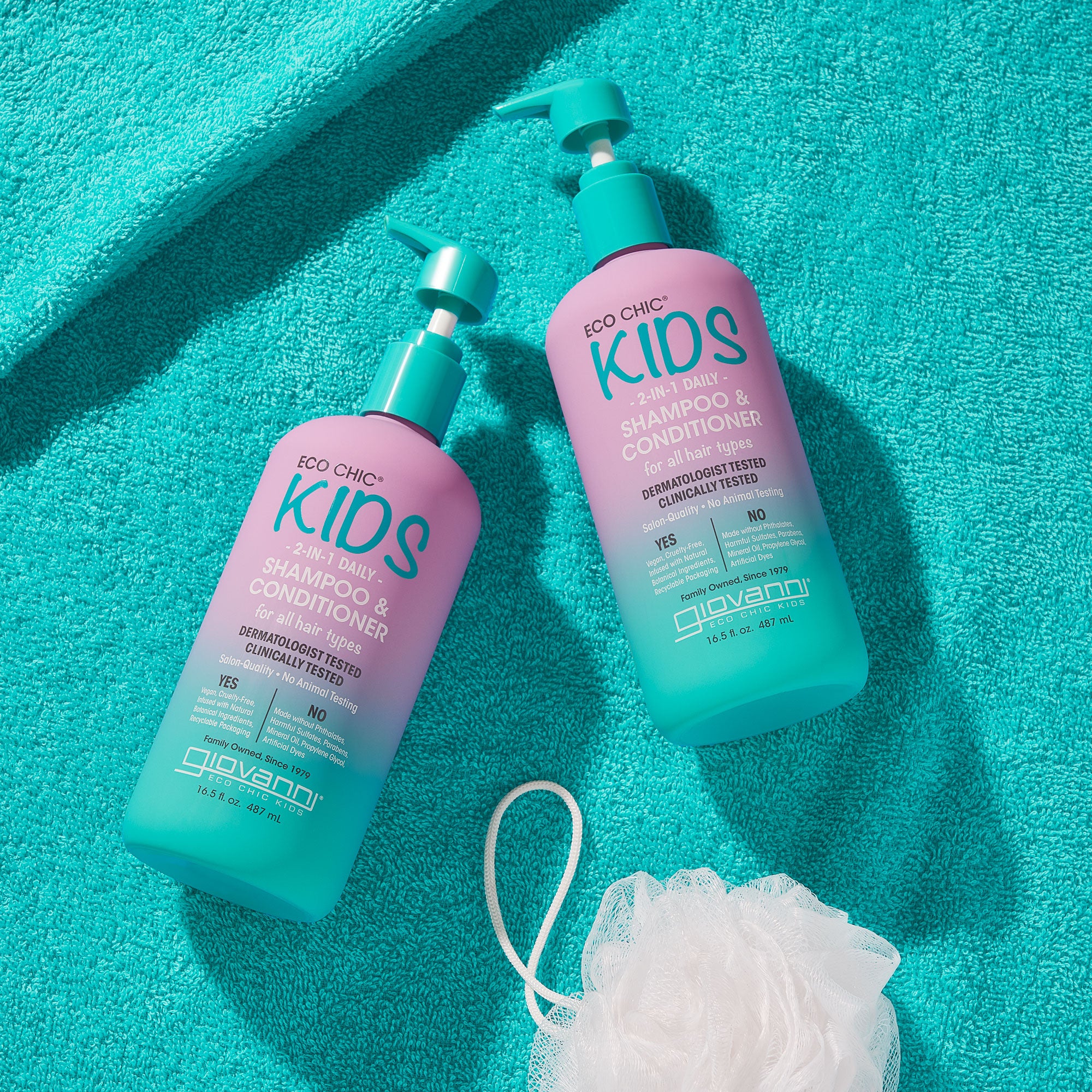 Two blue and purple pump bottles of Giovanni Cosmetics ECO CHIC® KIDS 2 IN 1 SHAMPOO & CONDITIONER sit on a turquoise towel next to a white bath pouf, featuring fun, colorful children’s haircare labels.