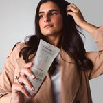 A woman with long dark hair, dressed in a beige trench coat and white shirt, poses against a plain light background holding Giovanni Cosmetics' L.A. HOLD™ STYLING GEL while touching her hair.