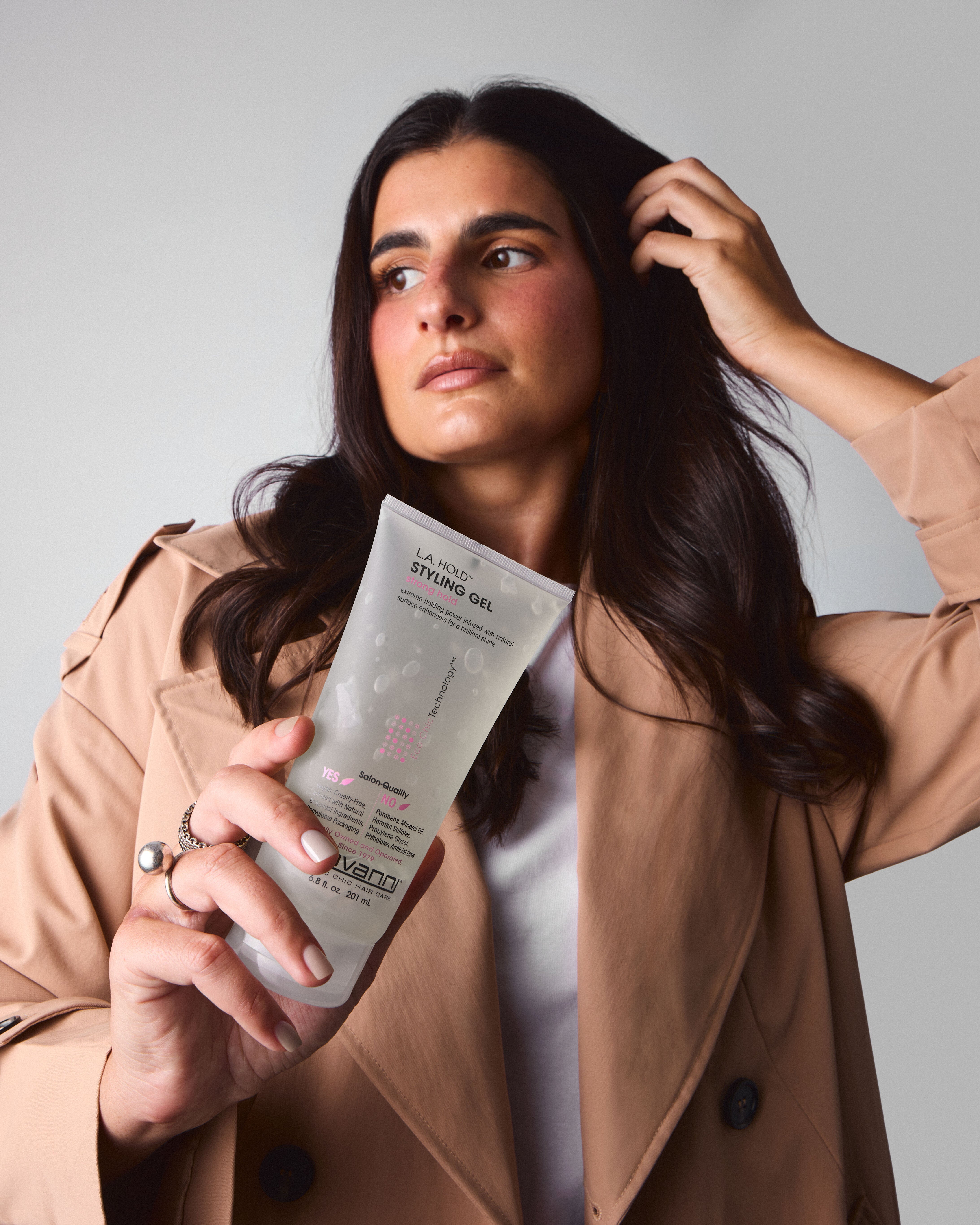 A woman with long dark hair, dressed in a beige trench coat and white shirt, poses against a plain light background holding Giovanni Cosmetics' L.A. HOLD™ STYLING GEL while touching her hair.