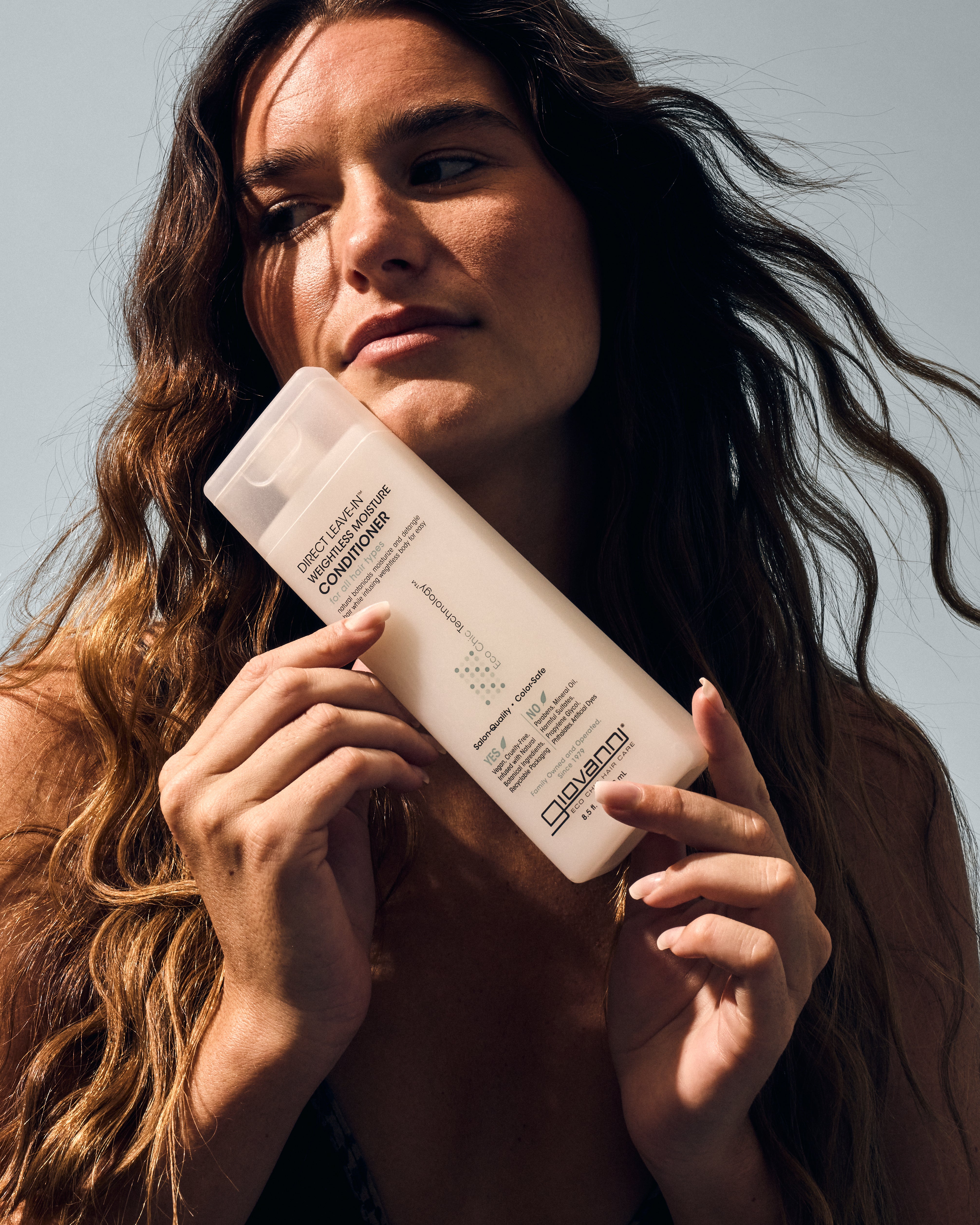 A woman with long wavy hair holds a bottle of Giovanni Cosmetics DIRECT LEAVE-IN™ WEIGHTLESS MOISTURE CONDITIONER close to her face, highlighting its weightless moisture benefits while looking away from the camera against a neutral background.
