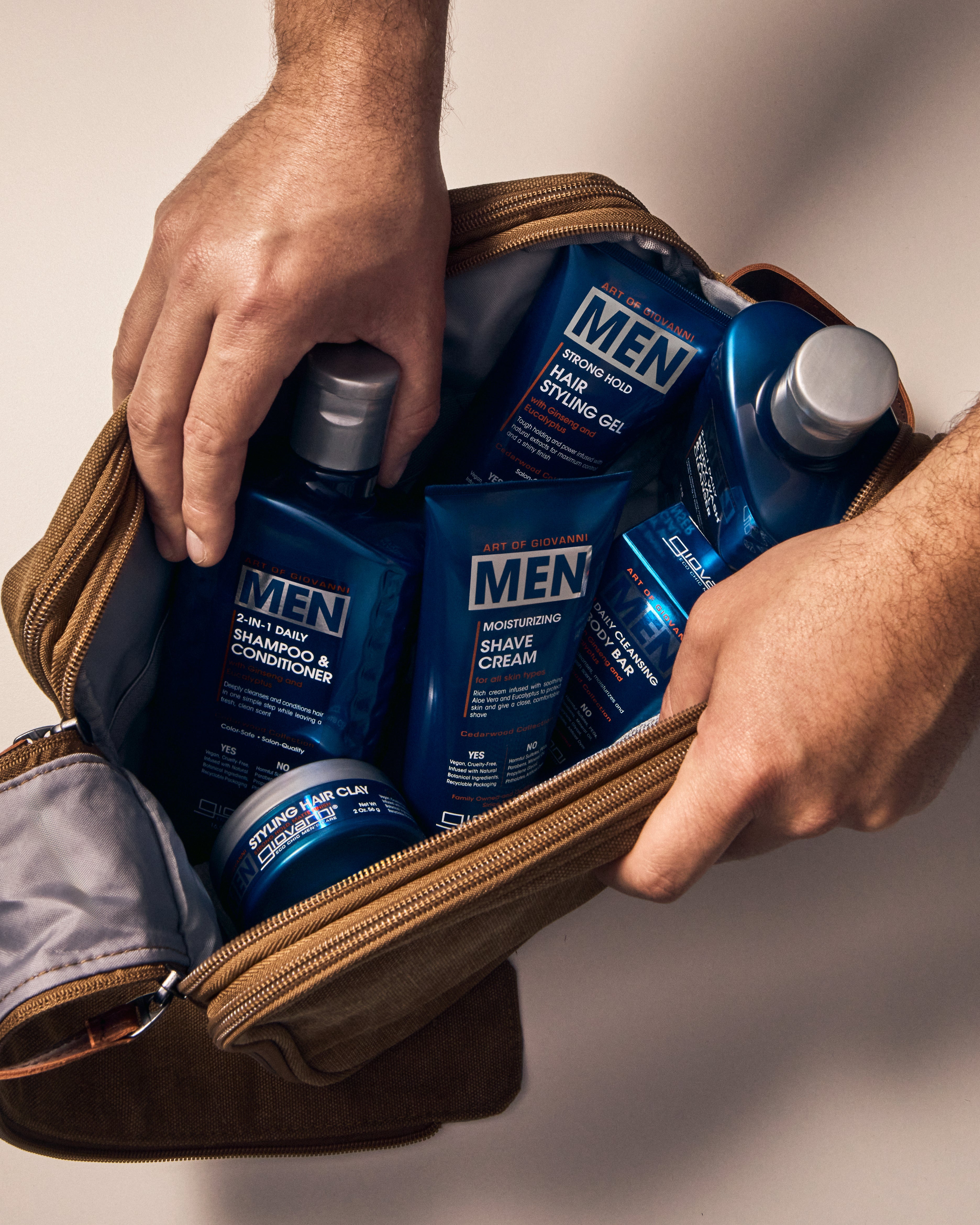 Someone places several blue bottles of Giovanni Cosmetics MEN 2-in-1 Daily Shampoo & Conditioner with Ginseng & Eucalyptus into a brown toiletry bag on a light surface.