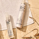 Image of Smooth As Silk Hair Detangling Spray and Frizz Be Gone Hair Serum laying on a towel on top of bathroom vanity tile. 