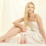 A woman in a strapless white dress with long blonde hair sits on the floor, gazing at the camera. Beside her are two bottles of Giovanni Cosmetics 2chic® FRIZZ BE GONE™ SHAMPOO, emphasizing her soft waves in a gentle, light setting.