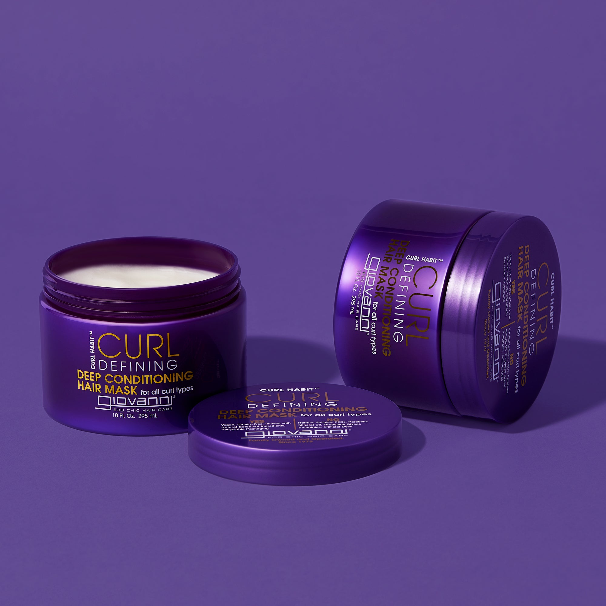 Three purple containers of Giovanni Cosmetics CURL HABIT™ Curl Defining Deep Conditioning Hair Mask are on a purple surface. One jar is open, revealing the curl hydration cream, with another closed jar and a lid beside it.