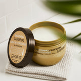 An open jar of Giovanni Cosmetics SMOOTHING CASTOR OIL LEAVE-IN CONDITIONER, a creamy, frizz-fighting formula, sits on a folded beige towel atop a white bathroom surface with its gold lid placed nearby.