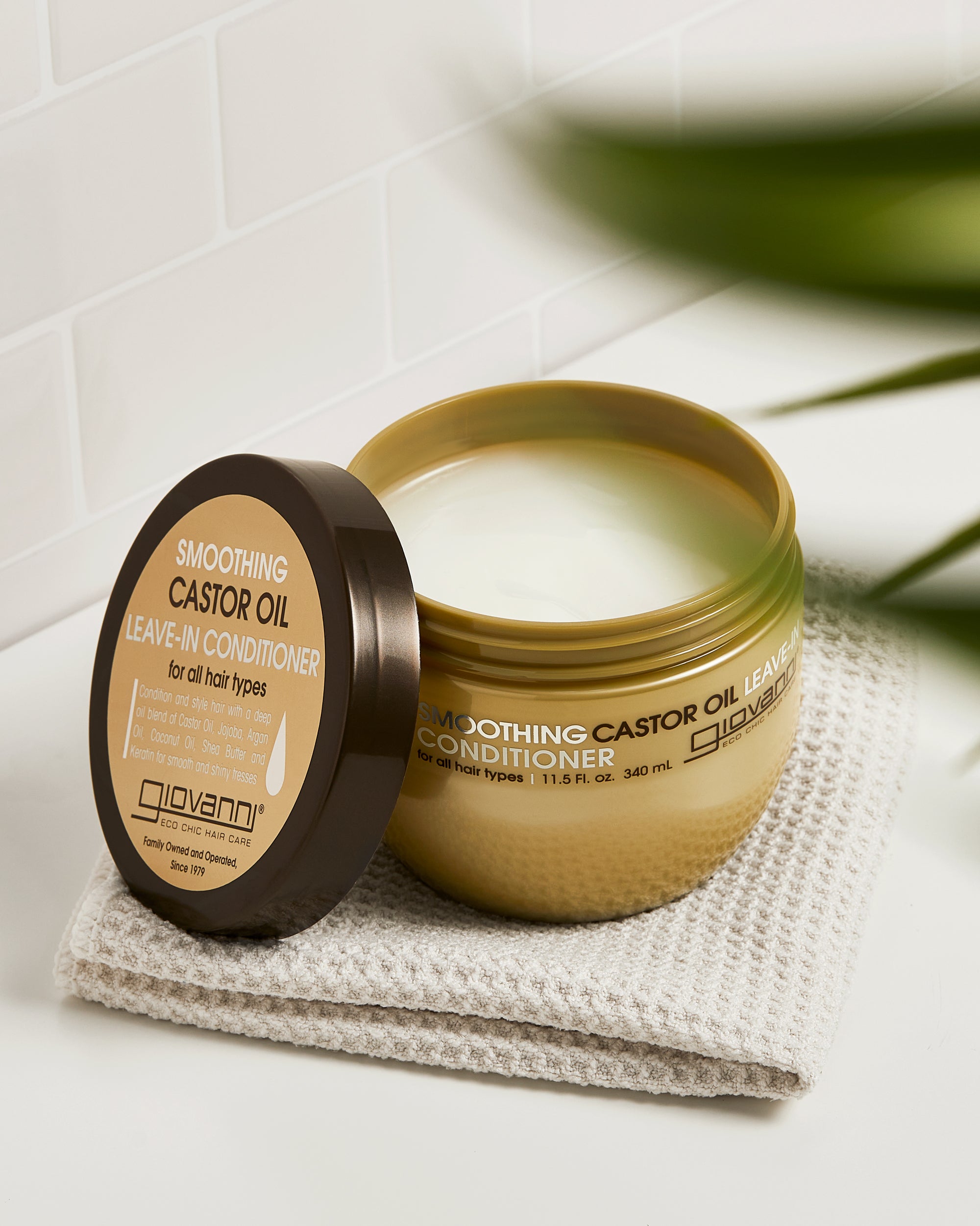 An open jar of Giovanni Cosmetics SMOOTHING CASTOR OIL LEAVE-IN CONDITIONER, a creamy, frizz-fighting formula, sits on a folded beige towel atop a white bathroom surface with its gold lid placed nearby.