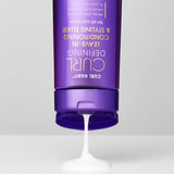 A purple bottle of Giovanni Cosmetics' CURL HABIT™ Curl Defining Leave-In Conditioning & Styling Elixir is upside down, dispensing its white, moisturizing cream onto a flat surface.
