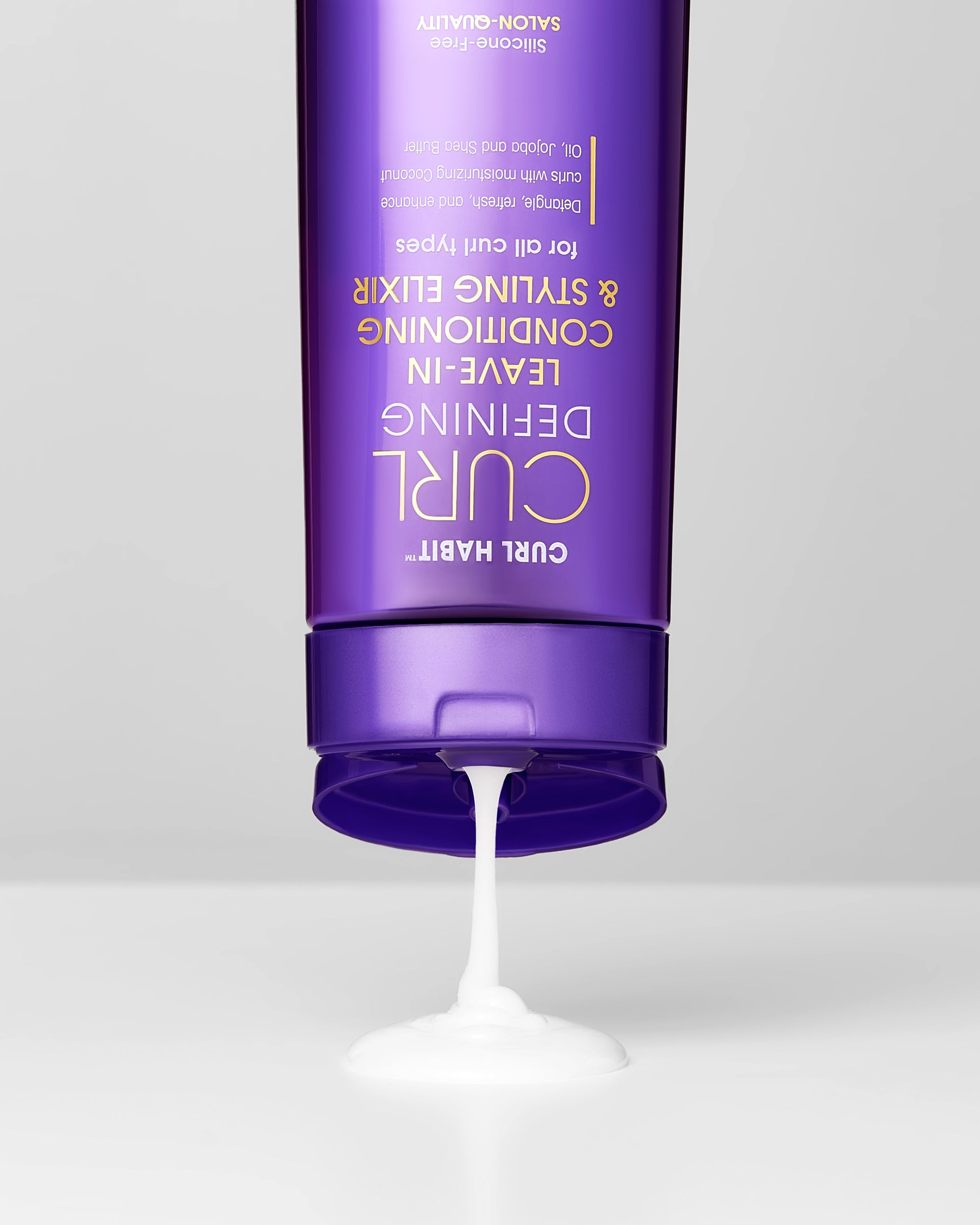 A purple bottle of Giovanni Cosmetics' CURL HABIT™ Curl Defining Leave-In Conditioning & Styling Elixir is upside down, dispensing its white, moisturizing cream onto a flat surface.