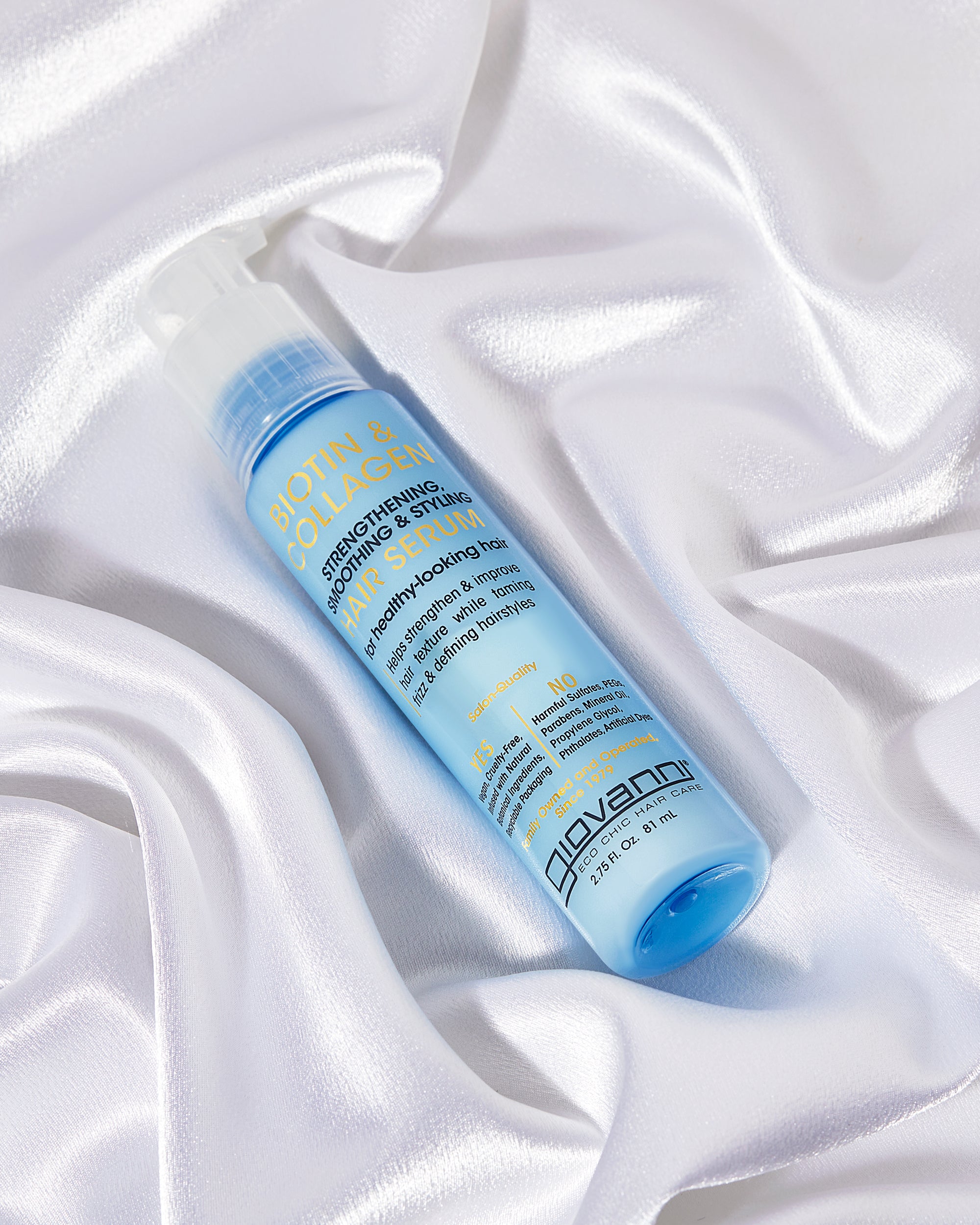 A blue bottle of Giovanni Cosmetics BIOTIN & COLLAGEN STRENGTHENING SMOOTHING & STYLING HAIR SERUM with a pump dispenser rests on white satin, featuring yellow and blue label text for boosting hair volume.