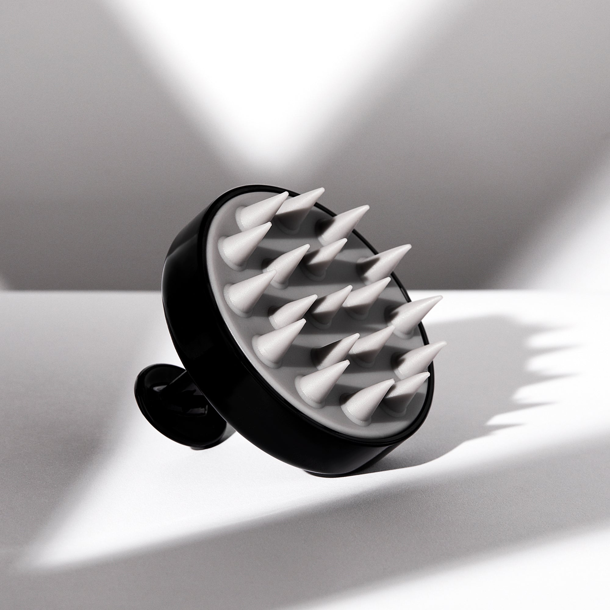 The Giovanni Cosmetics SCALP MASSAGER & SHAMPOO BRUSH (Black Handle) features pointed silicone bristles and sits on a white surface, casting geometric shadows—a salon-quality tool ideal for natural hair care.