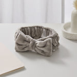 The Giovanni Cosmetics PLUSH MULTI-PURPOSE SPA HEADBAND in gray, featuring a large bow and non-slip elastic, sits on a white surface near a notebook—ideal for enhancing your beauty routine.