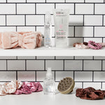 Two white shelves against subway tile hold hair care products, pink and brown scrunchies, the PLUSH MULTI-PURPOSE SPA HEADBAND (blush), a silicone scalp massager, plus Giovanni Cosmetics styling gel and anti-frizz serum bottles.