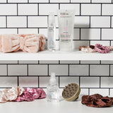 Two white shelves against subway tile hold hair care products, pink and brown scrunchies, the PLUSH MULTI-PURPOSE SPA HEADBAND (blush), a silicone scalp massager, plus Giovanni Cosmetics styling gel and anti-frizz serum bottles.
