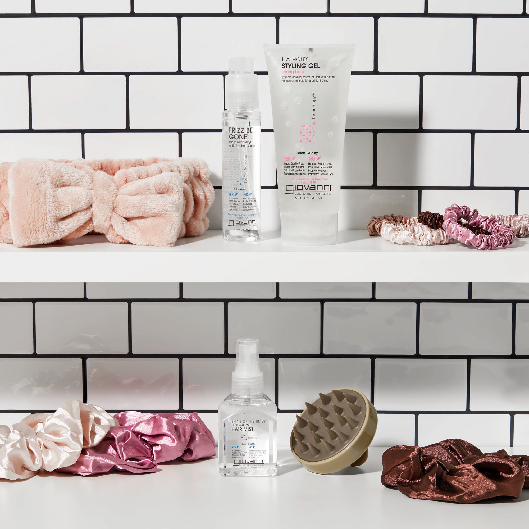 Two white shelves against subway tile hold hair care products, pink and brown scrunchies, the PLUSH MULTI-PURPOSE SPA HEADBAND (blush), a silicone scalp massager, plus Giovanni Cosmetics styling gel and anti-frizz serum bottles.