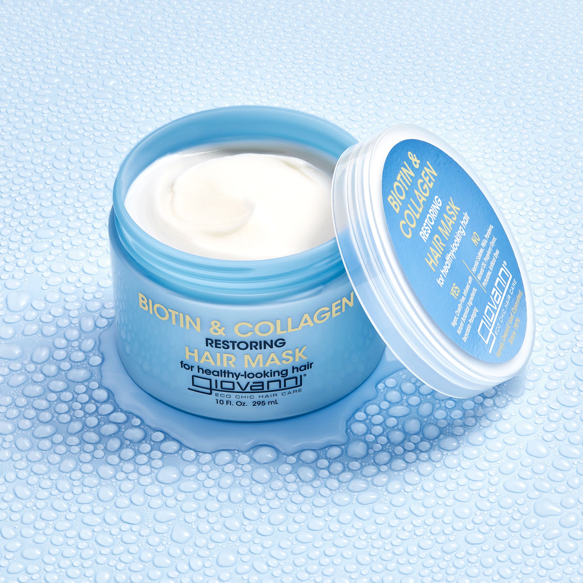 A blue jar of Giovanni Cosmetics BIOTIN & COLLAGEN RESTORING HAIR MASK sits open on a wet surface, revealing its creamy white formula—ideal for adding volume to your hair.