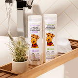 Two bottles of Giovanni Cosmetics PROFESSIONAL 2-IN-1 PET SHAMPOO & CONDITIONER - TEA TREE & ALOE rest on a wooden bath tray beside a potted plant and white towel, creating an inviting pet care scene against a white tiled wall.