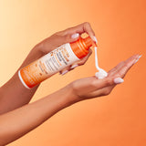 A person dispenses 2chic® ULTRA-VOLUME FOAM STYLING MOUSSE by Giovanni Cosmetics from an orange and white bottle onto their palm, set against an orange background.
