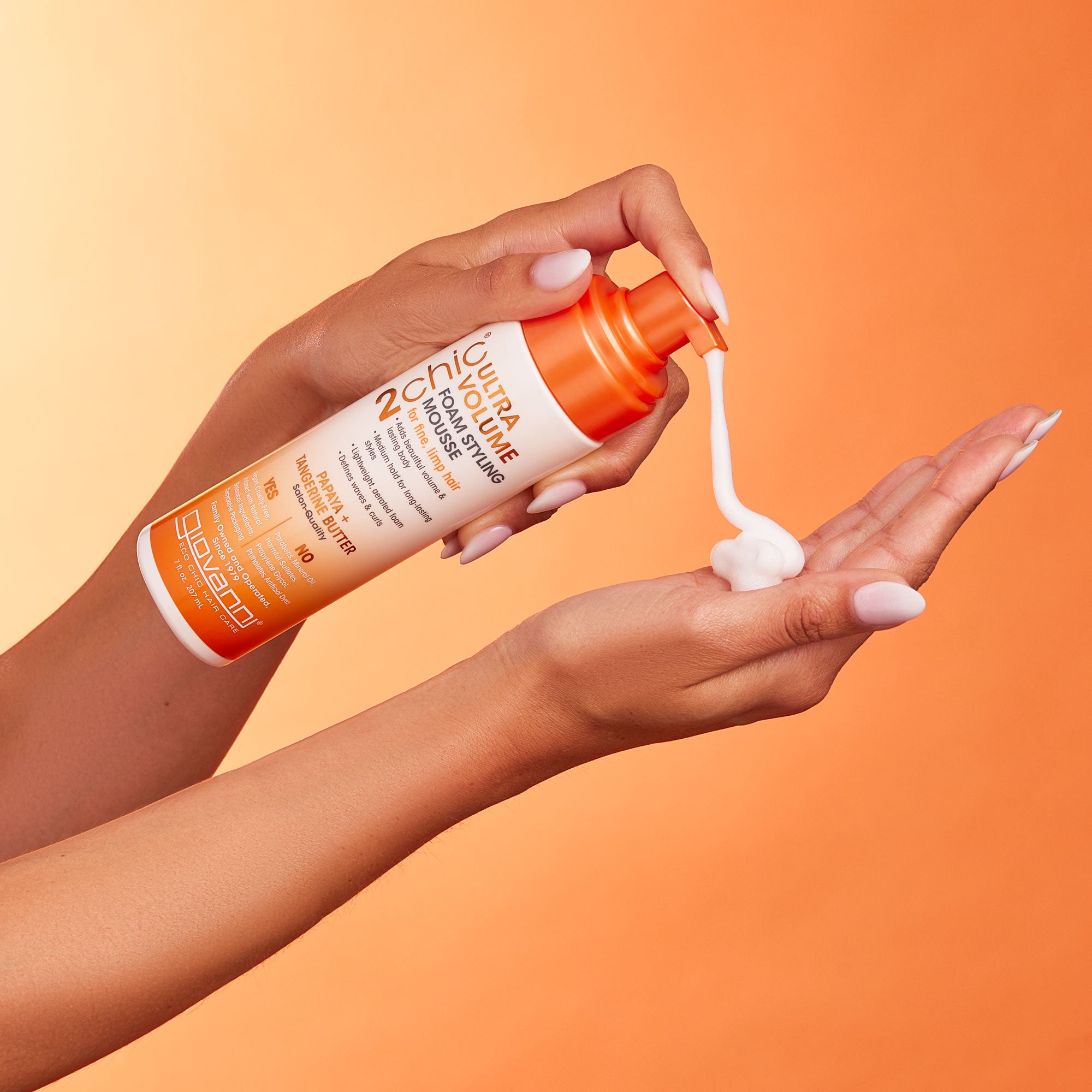 A person dispenses 2chic® ULTRA-VOLUME FOAM STYLING MOUSSE by Giovanni Cosmetics from an orange and white bottle onto their palm, set against an orange background.