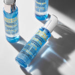 Three blue bottles of Giovanni Cosmetics BIOTIN & COLLAGEN REBONDING & RESTORING HAIR OIL are displayed on a light surface—one lying down and two standing upright—highlighting their volumizing formula and soft shadows.