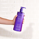 A hand holds a purple bottle of Giovanni Cosmetics CURL HABIT™ CURL DEFINING SHAMPOO, a curl shampoo that defines and moisturizes curly hair, in front of a white tile wall with water droplets in the air.