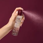 A hand sprays Giovanni Cosmetics 2chic® ULTRA-SLEEK FLAT IRON STYLING MIST against a burgundy background, releasing a fine mist.