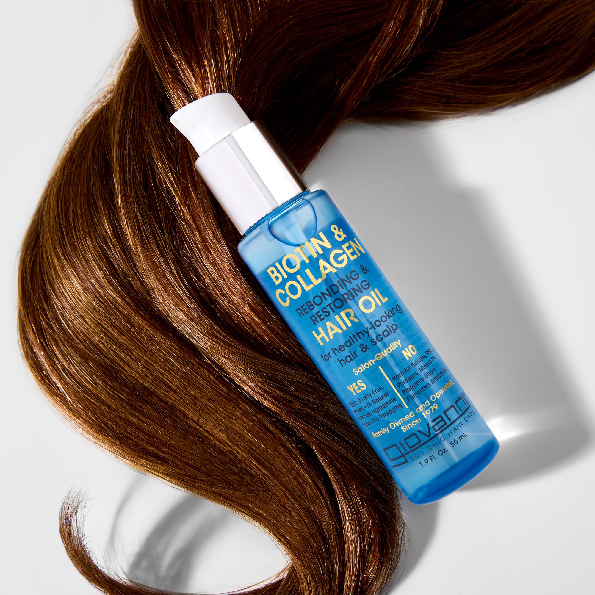 A blue bottle of Giovanni Cosmetics BIOTIN & COLLAGEN REBONDING & RESTORING HAIR OIL sits atop smooth, shiny brown hair. Its white pump and gold text showcase benefits for healthy, glossy hair and increased volume.