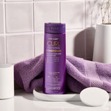 A purple bottle of Giovanni Cosmetics CURL HABIT™ CURL DEFINING CONDITIONER sits on a white tray beside a matching towel and container, all set against glossy white tiled walls.