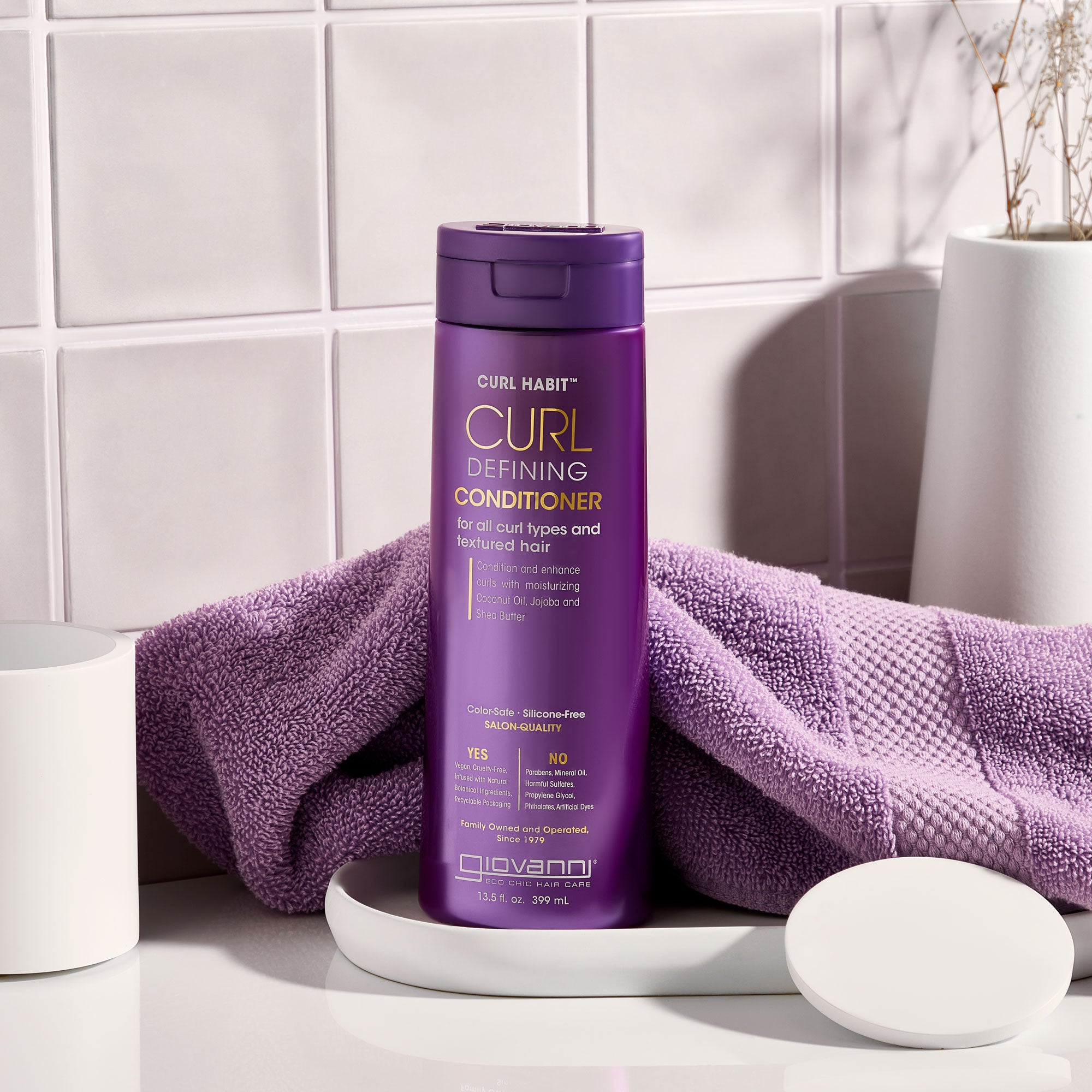 A purple bottle of Giovanni Cosmetics CURL HABIT™ CURL DEFINING CONDITIONER sits on a white tray beside a matching towel and container, all set against glossy white tiled walls.