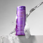 A purple bottle of Giovanni Cosmetics CURL HABIT™ CURL DEFINING SHAMPOO stands upright with water splashing around it and soap suds at the base, set against a light gray background.