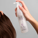 A hand holds a bottle of Giovanni Cosmetics ROOT 66™ MAX VOLUME DIRECTIONAL HAIR ROOT LIFTING SPRAY near brown hair. This color-safe product features printed text on a translucent bottle with a convenient pump nozzle, ready for use.