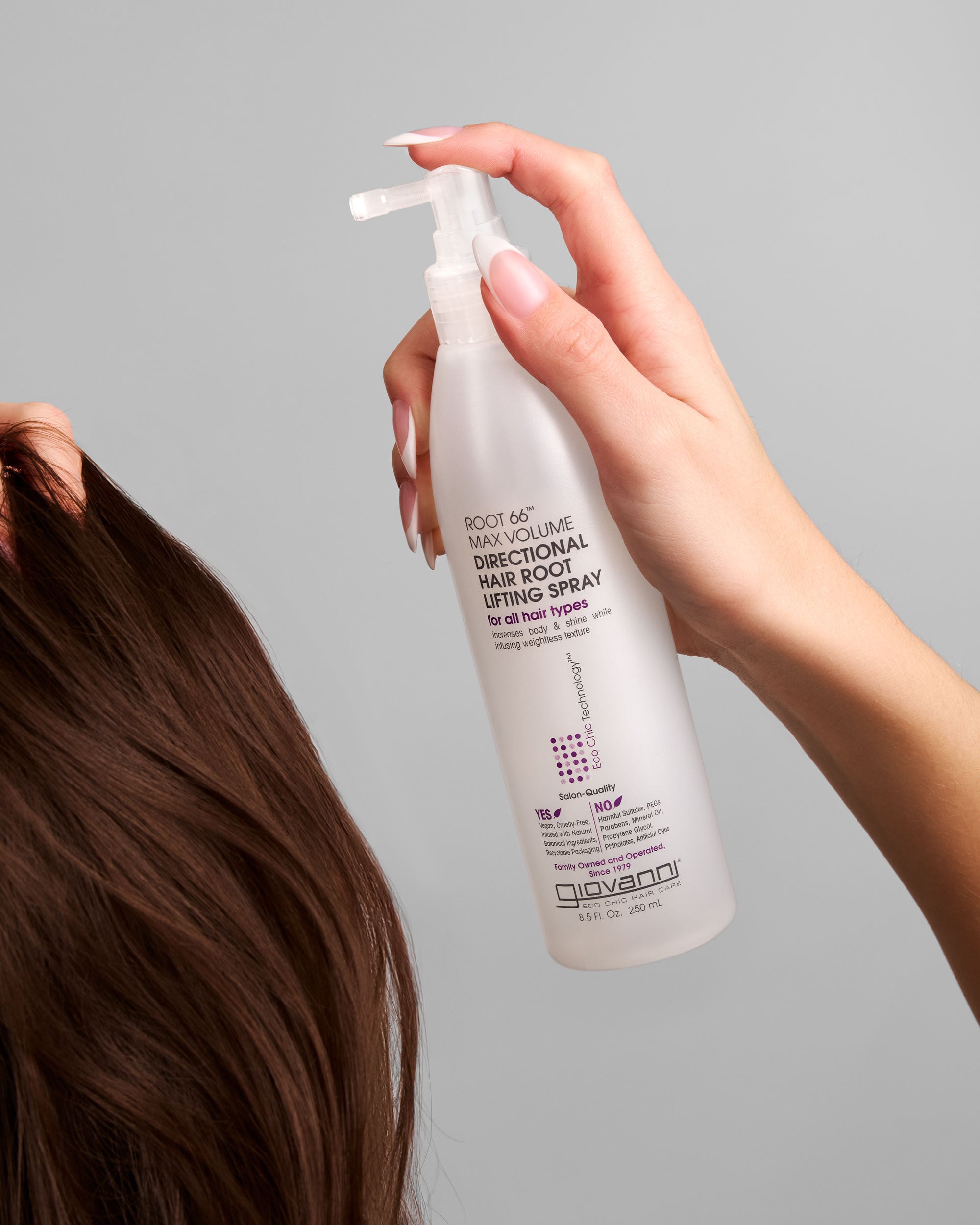 A hand holds a bottle of Giovanni Cosmetics ROOT 66™ MAX VOLUME DIRECTIONAL HAIR ROOT LIFTING SPRAY near brown hair. This color-safe product features printed text on a translucent bottle with a convenient pump nozzle, ready for use.