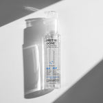 A clear bottle of Giovanni Cosmetics FRIZZ BE GONE™ SUPER-SMOOTHING, ANTI-FRIZZ HAIR SERUM stands upright in bright sunlight, casting sharp shadows on a white and gray background. The front label highlights its anti-frizz and smoothing benefits.