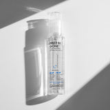 A clear bottle of Giovanni Cosmetics FRIZZ BE GONE™ SUPER-SMOOTHING, ANTI-FRIZZ HAIR SERUM stands upright in bright sunlight, casting sharp shadows on a white and gray background. The front label highlights its anti-frizz and smoothing benefits.