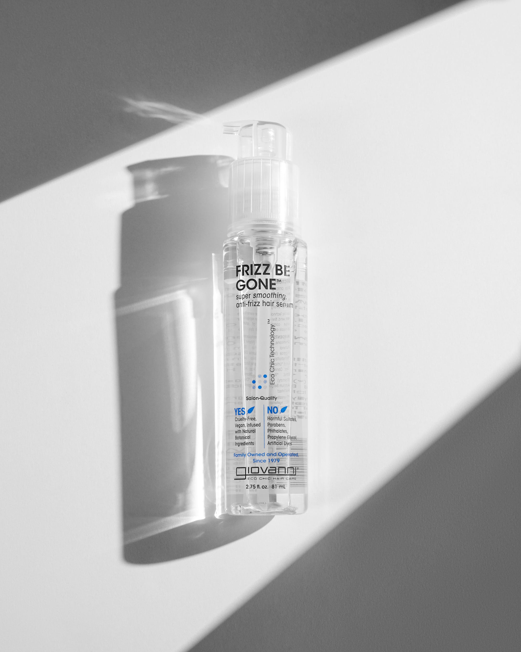 A clear bottle of Giovanni Cosmetics FRIZZ BE GONE™ SUPER-SMOOTHING, ANTI-FRIZZ HAIR SERUM stands upright in bright sunlight, casting sharp shadows on a white and gray background. The front label highlights its anti-frizz and smoothing benefits.