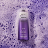 A bottle of Giovanni Cosmetics COLOR CHIC™ PURPLE SHAMPOO for color-treated, lightened hair sits on purple foam. The white-to-purple bottle helps reduce brassy tones in blonde hair.
