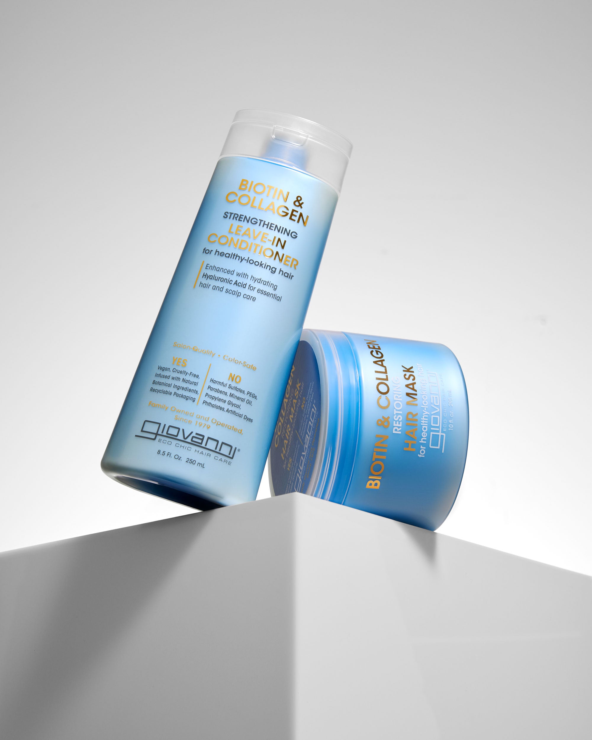 Two blue Giovanni Cosmetics BIOTIN & COLLAGEN RESTORING HAIR MASKs for hair volume are displayed on a white geometric pedestal against a light gray background.