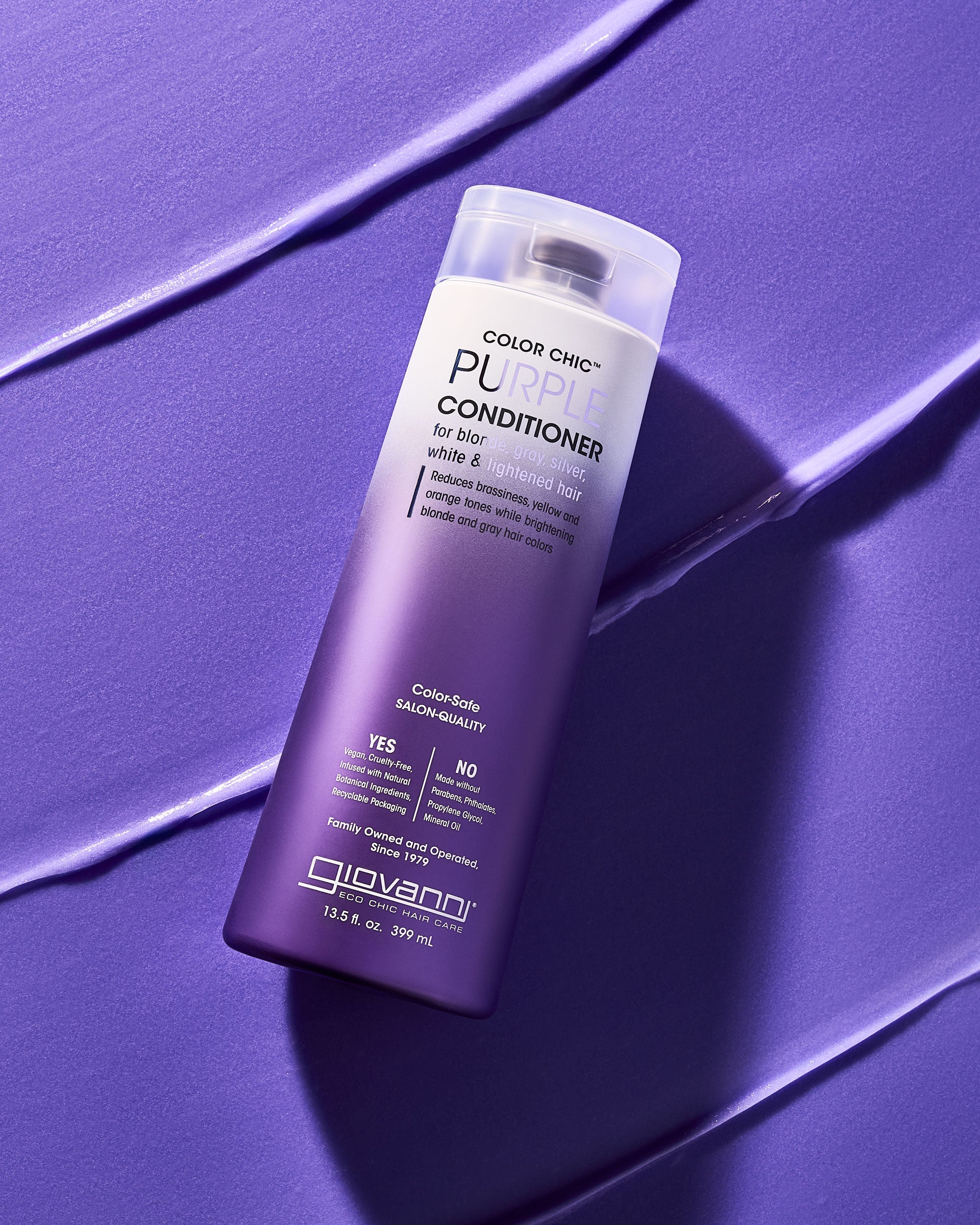 A bottle of Giovanni Cosmetics COLOR CHIC™ PURPLE CONDITIONER for blonde or silver hair stands upright on a textured purple surface, with matching conditioner streaks nearby and the label clearly visible.