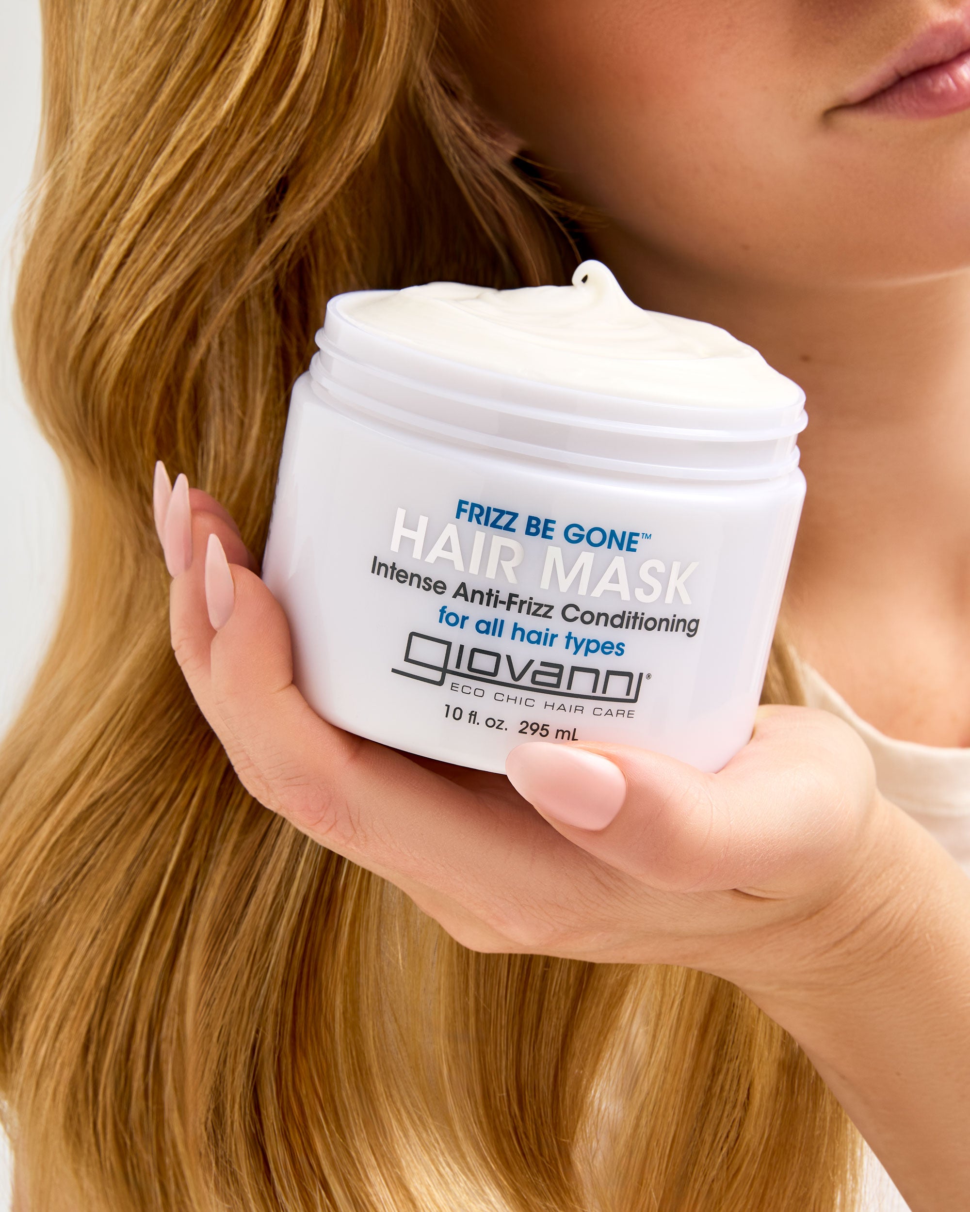 Hand holding a jar of Giovanni Frizz Be Gone hair mask with blonde hair in the background