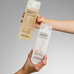 Two hands hold Giovanni Cosmetics hair care bottles against a gray background—one is 50:50 Balanced Hydrating-Clarifying Shampoo, the other is 50:50 BALANCED™ HYDRATING-CALMING CONDITIONER.